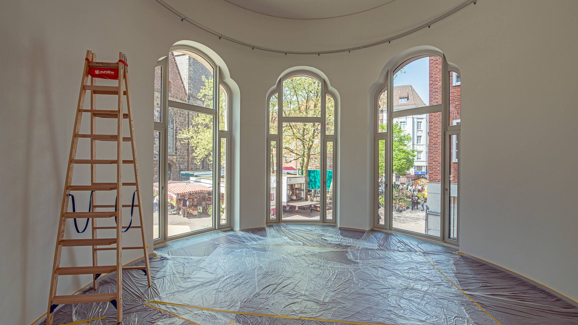 Blick in den runden Raum, dessen Fenster zur Innenstadt zeigen. Eine Leiter steht im Raum und der Boden ist mit Folie abgedeckt.