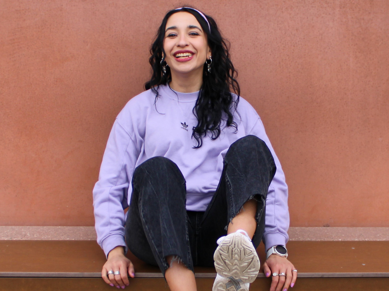 Charleen Hahn sitzt im lila Pullover lachend auf einer ockerfarbenen Mauer.