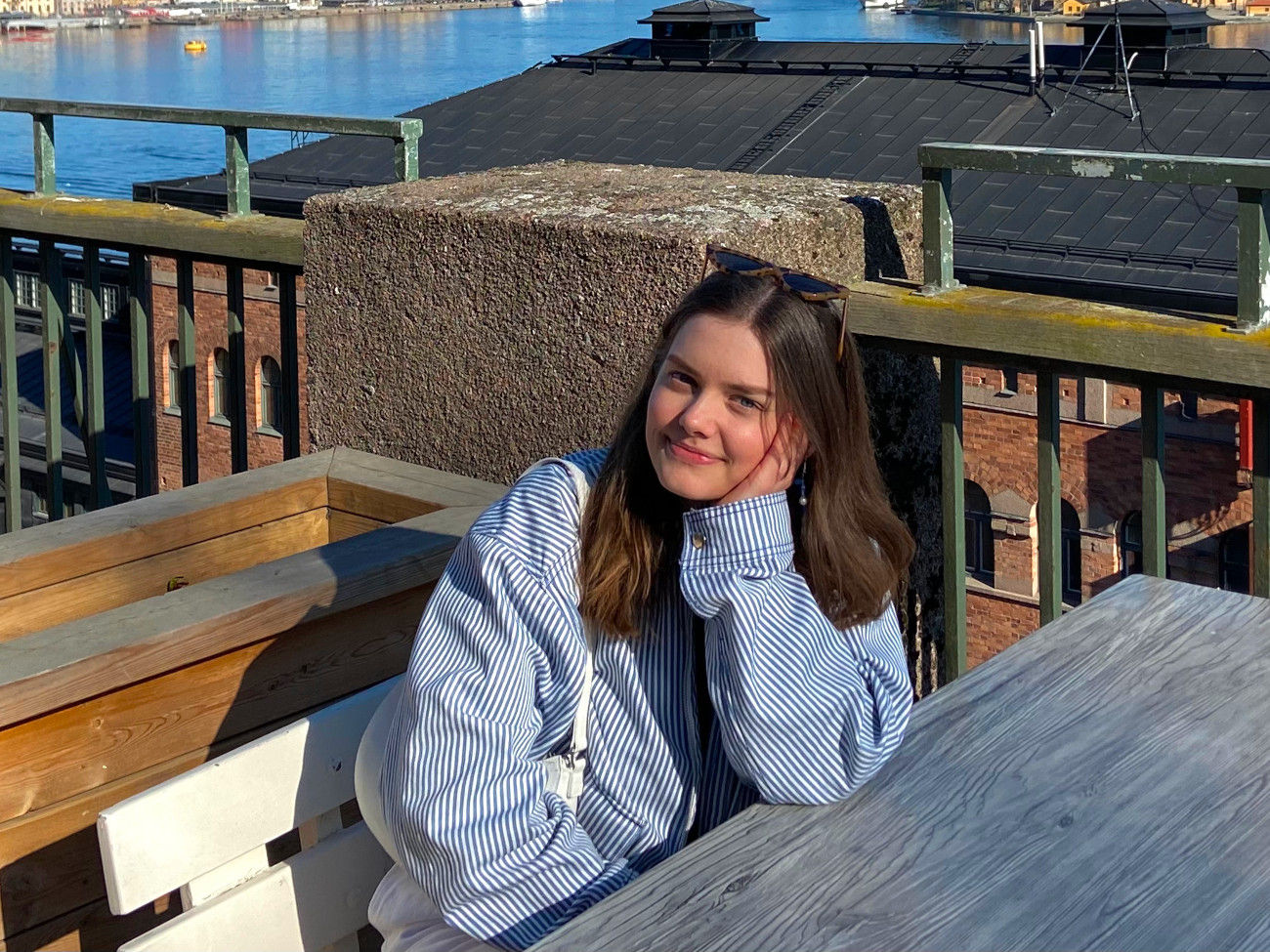 Hannah Bruns in blau-weiß gestreifter Jacke sitzt an einem Tisch auf einer Terrasse. Im Hintergrund ist ein Fluss zu sehen.