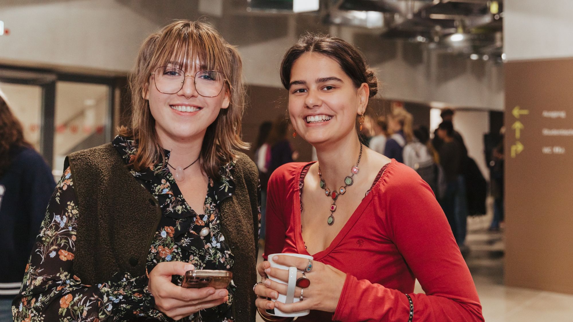Two women are smiling at the camera. One is holding a cup, the other a cell phone. 