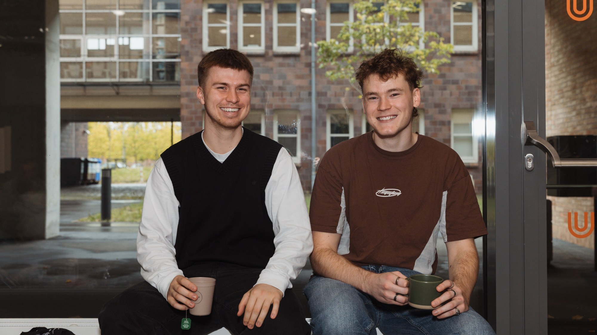 Two people are sitting together, smiling at the camera and each holding a cup in their hand. 