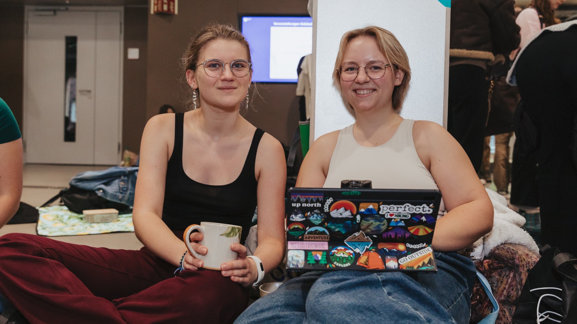 Two women are sitting together on the floor. One is holding a cup in her hand and the other has a laptop on her lap.