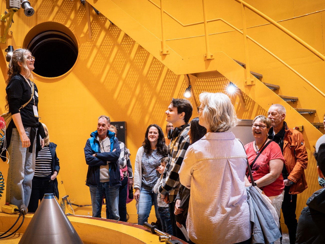 A group of people are standing in a yellow room, looking up at the campus tour guide. 