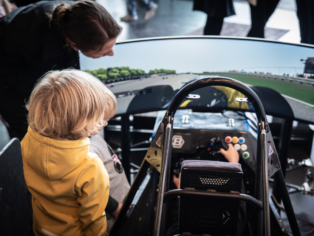 A person is sitting in a racing simulator, while an older person and another child watch. 