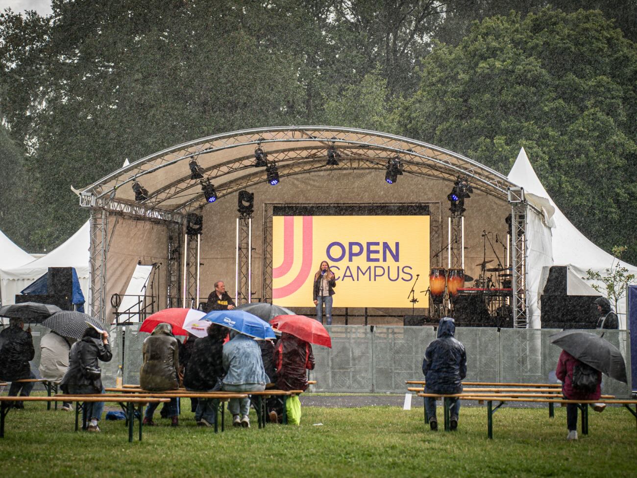 The guests are sitting in front of the Open Campus stage, holding umbrellas and wearing rain jackets, as it rains. 