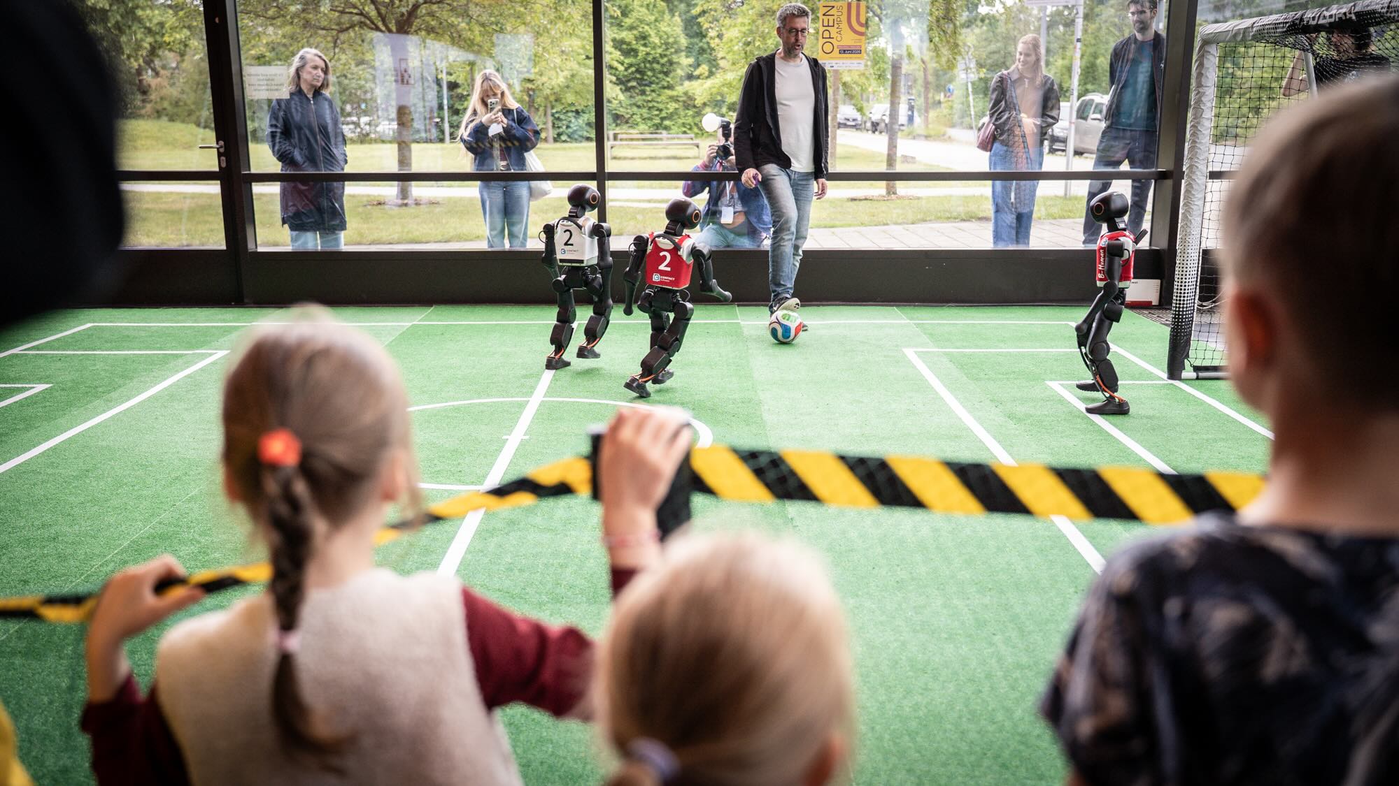 The picture shows a robot soccer tournament that children are watching. 