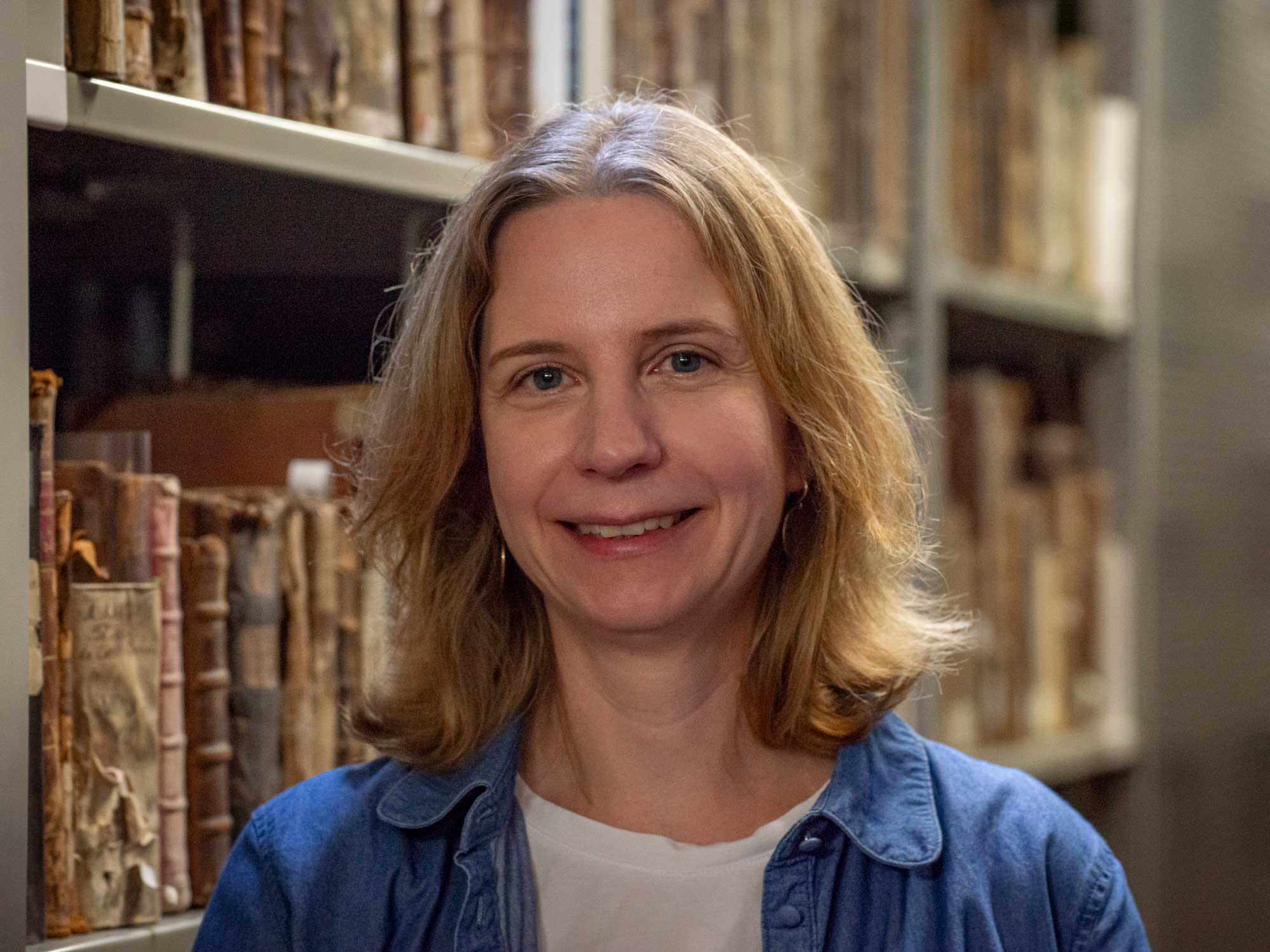 Im Vordergrund lächelt eine Frau. Im Hintergrund sieht man ein mit Büchern gefülltes Regal.