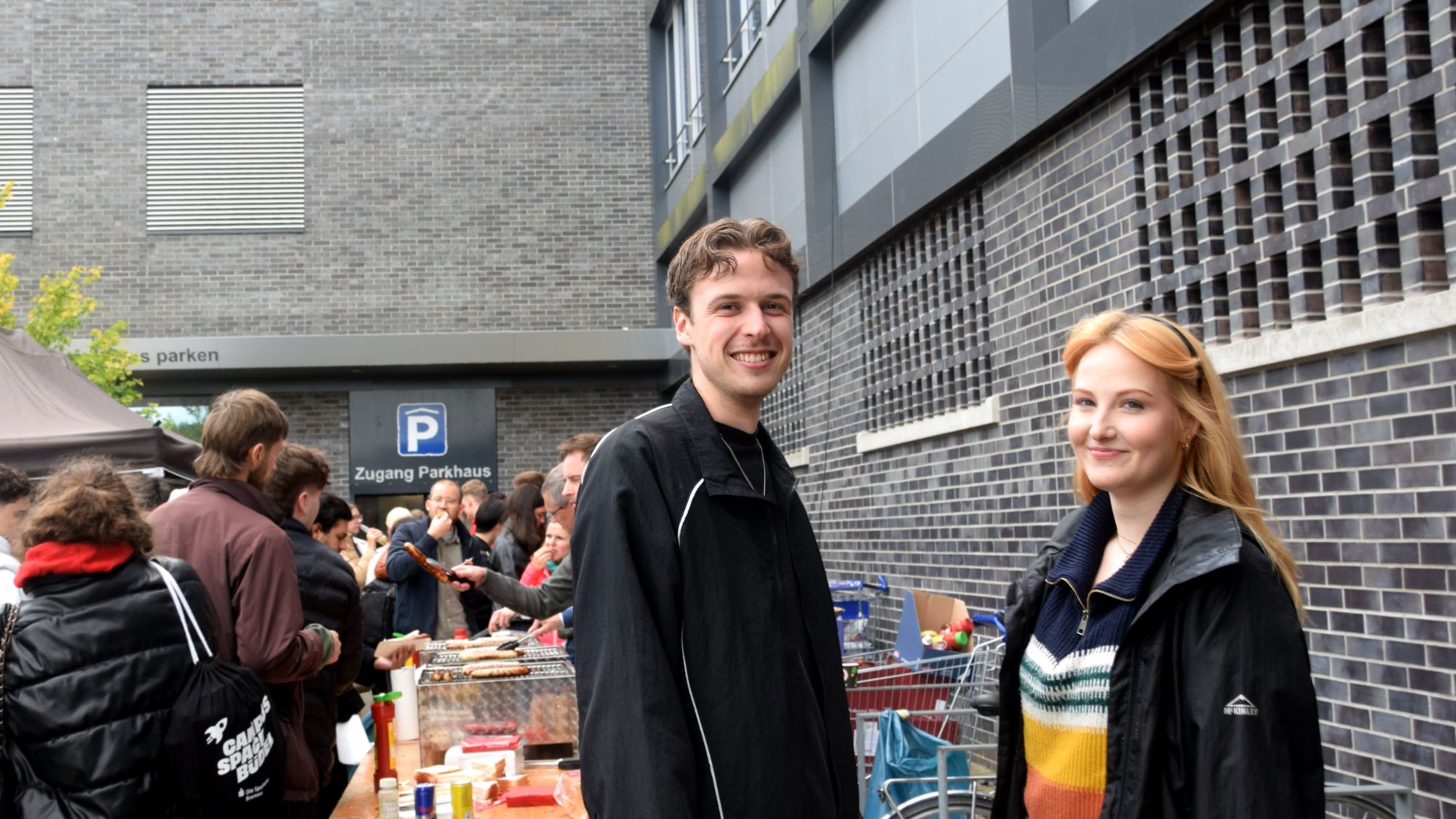 Two people are smiling at the camera. In the background, you can see a line of people waiting at the grills.