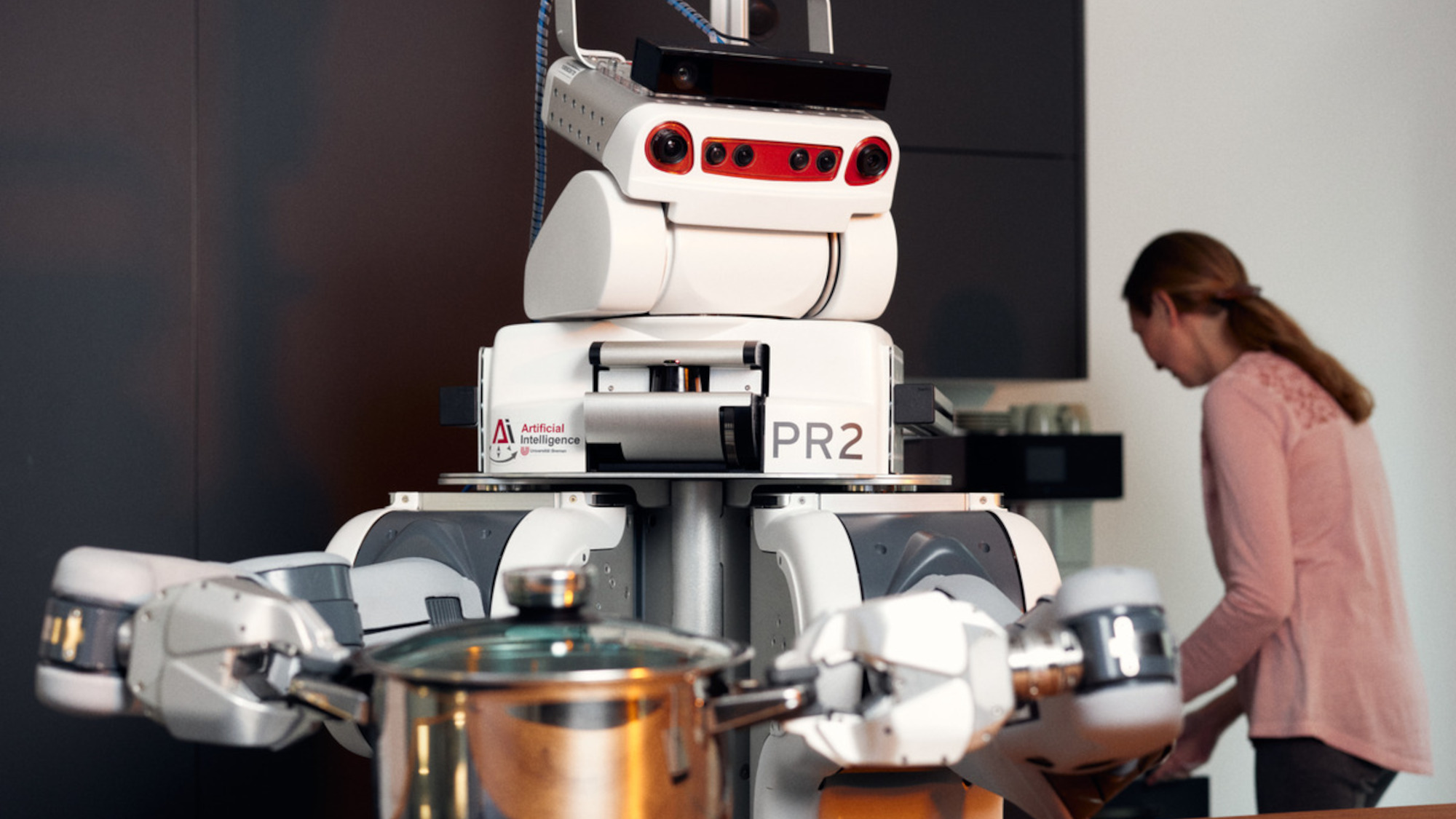 Ein Roboter hält einen Kochtopf in seinen Händen. Er hilft der Frau, die im Hintergrund zu sehen ist, beim Kochen.