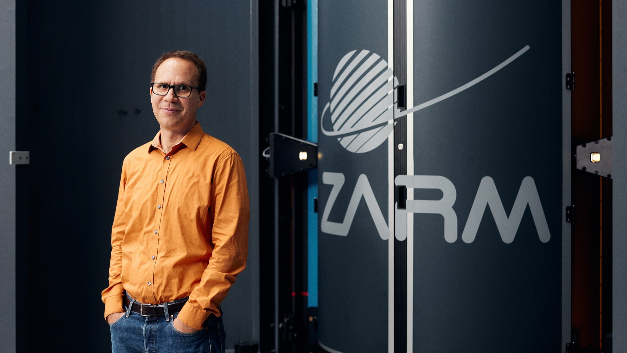 A man stands next to the ZARM logo and smiles into the camera.
