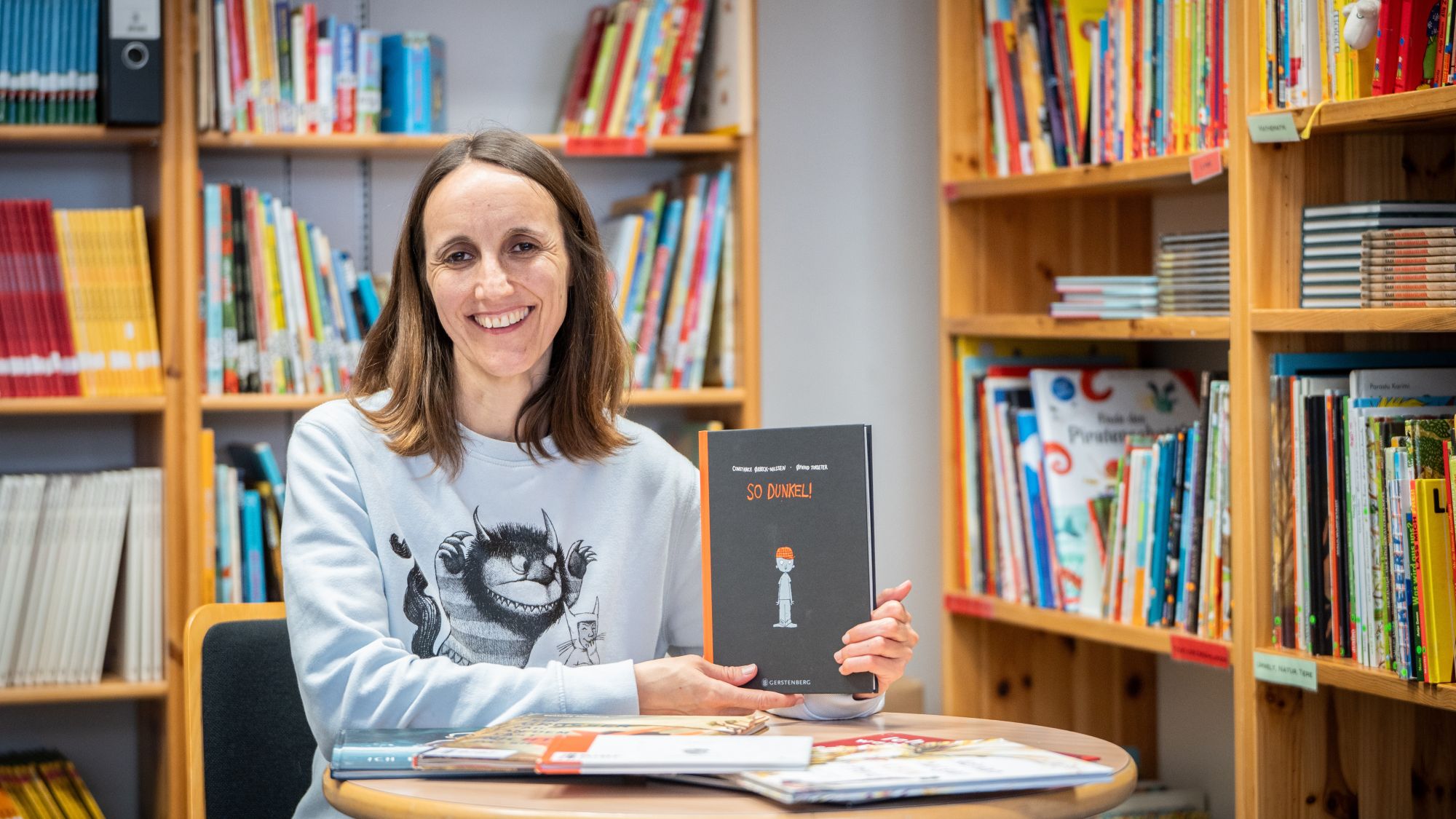 Elisabeth Hollerweger sitzt an einem Tisch in der Bilderbuchbibliothek und hält ein Buch mit dem Titel „So dunkel“ in den Händen.