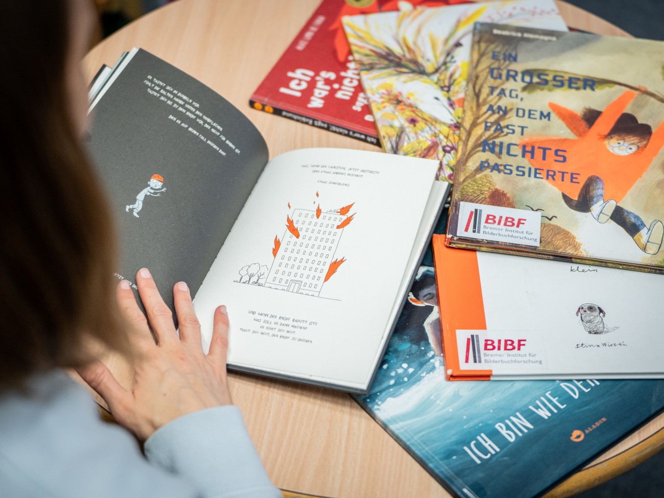 Blick über Elisabeth Hollerwegers Schulter, während sie in einem Bilderbuch blättert. Vor ihr liegen weitere Bilderbücher auf dem Tisch.