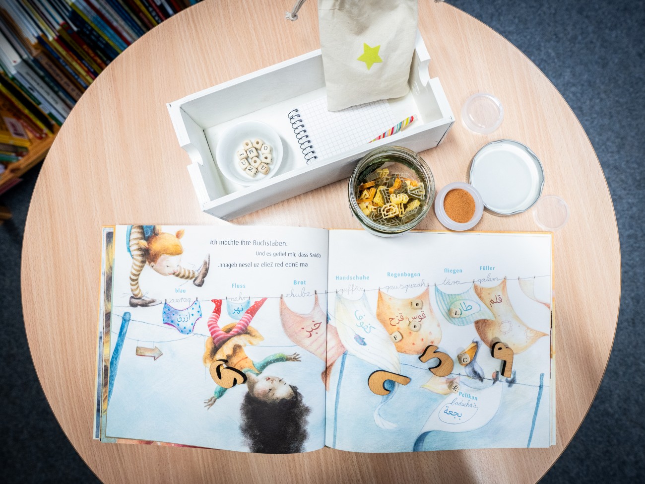 Man sieht einen Tisch von oben. Darauf liegen ein aufgeschlagenes Bilderbuch und kleine Behälter mit Buchstabennudeln, einem rötlichen Pulver und Schreibutensilien.