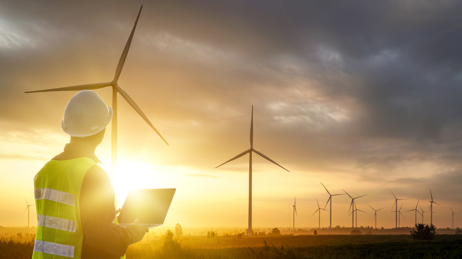 Mann mit Warnweste, Laptop und Helm blickt auf einen Windpark im Sonnenuntergang