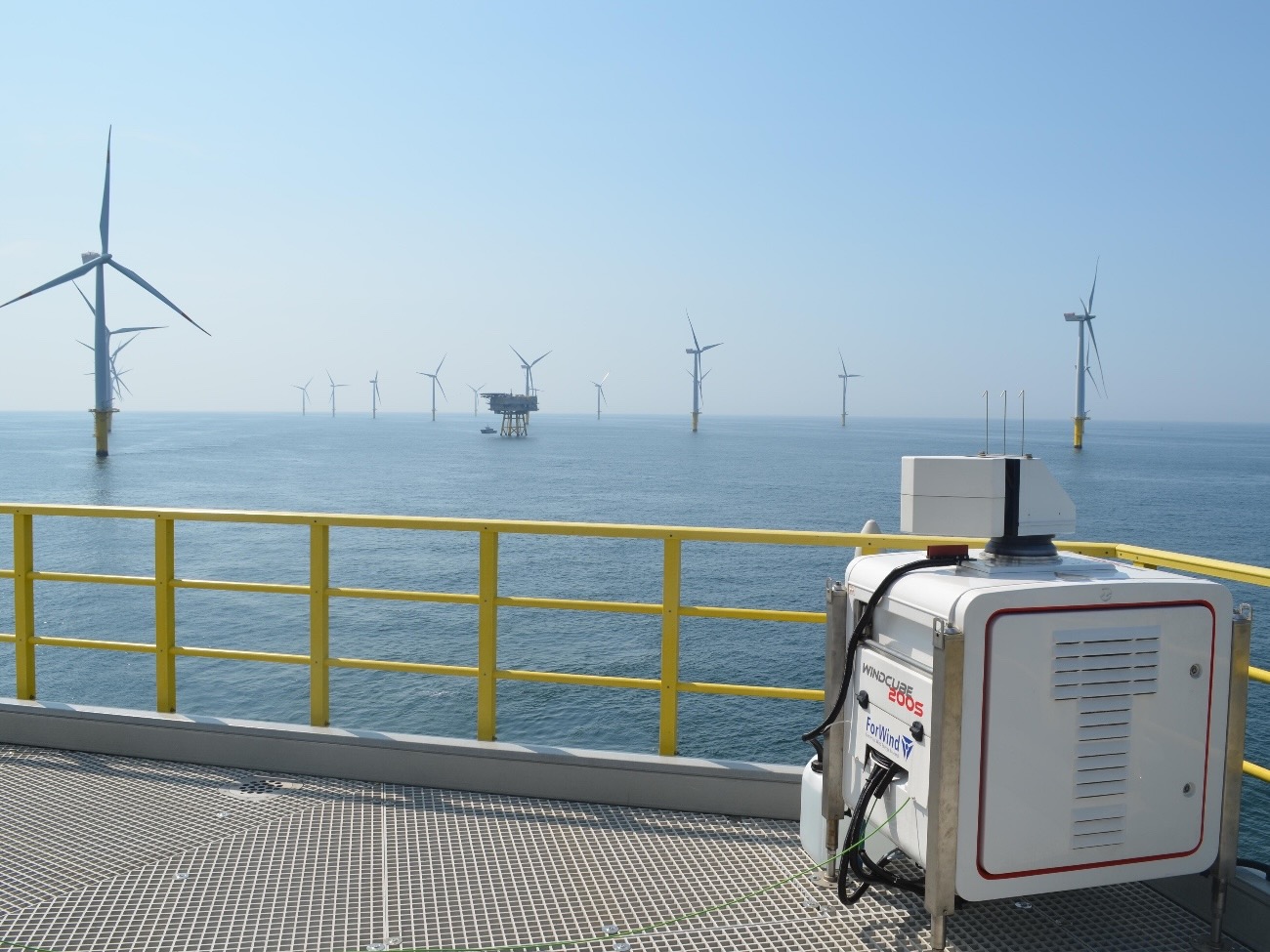 A photo of a box-shaped lidar device standing on a coastal platform. In the background, wind turbines rise out of the sea. 
