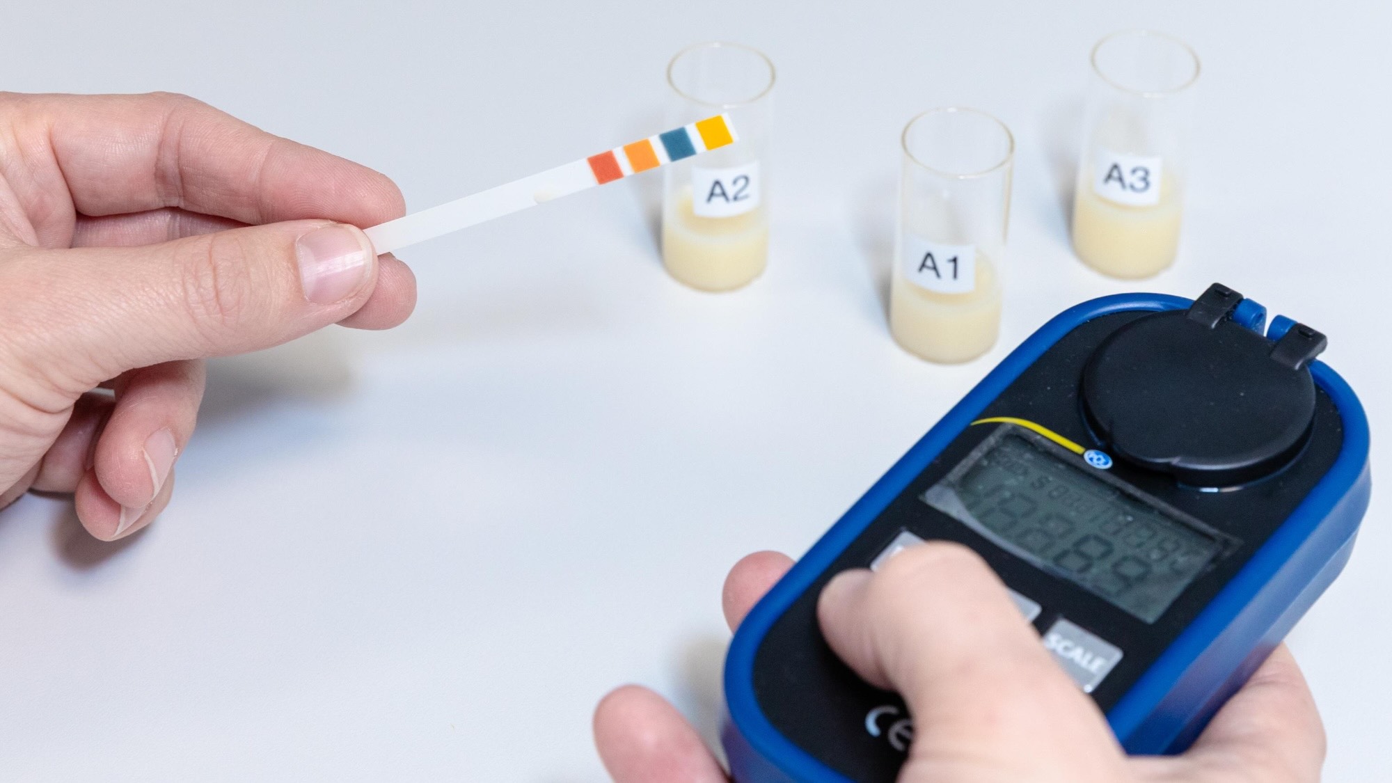 hands are visible. One is holding a digital measuring device and the other is holding a test strip. There are also three samples of manure in glass jars on the table.