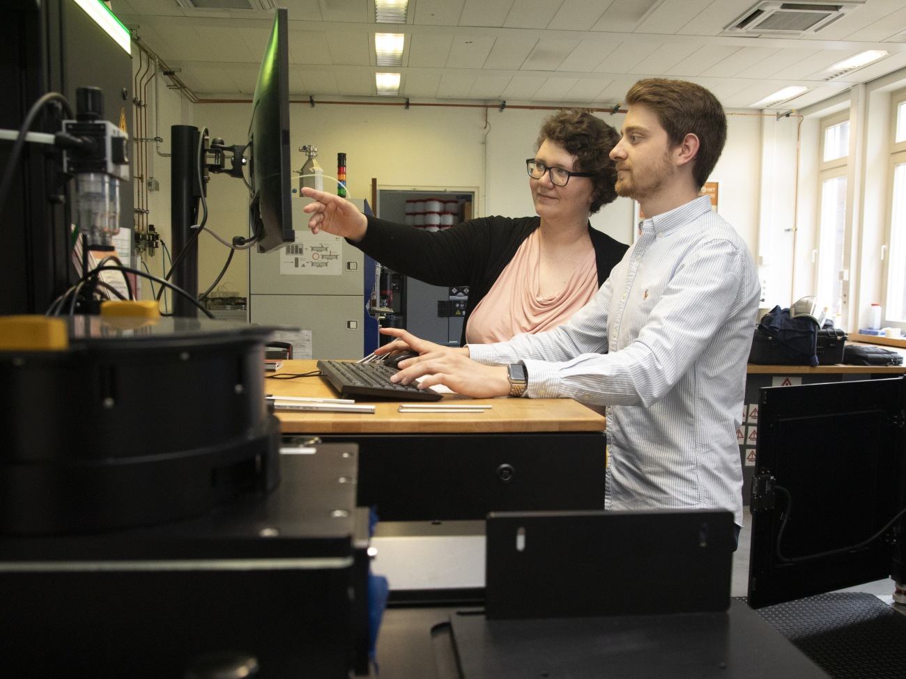Two people are standing at a standing desk and are looking at a monitor. The woman is pointing at it.