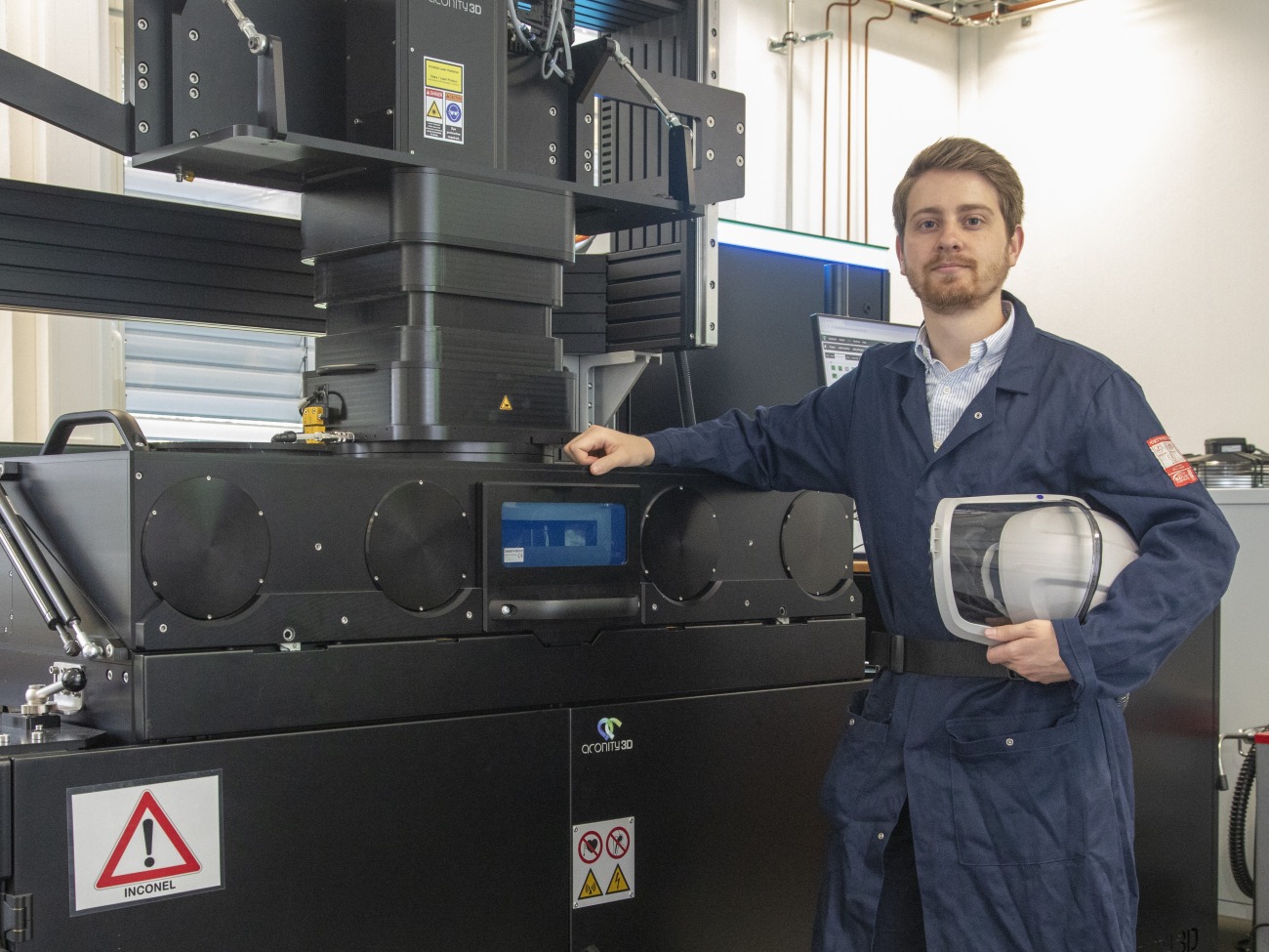 A man in a blue collar stands in front of a machine with a safety helmet under his arm.