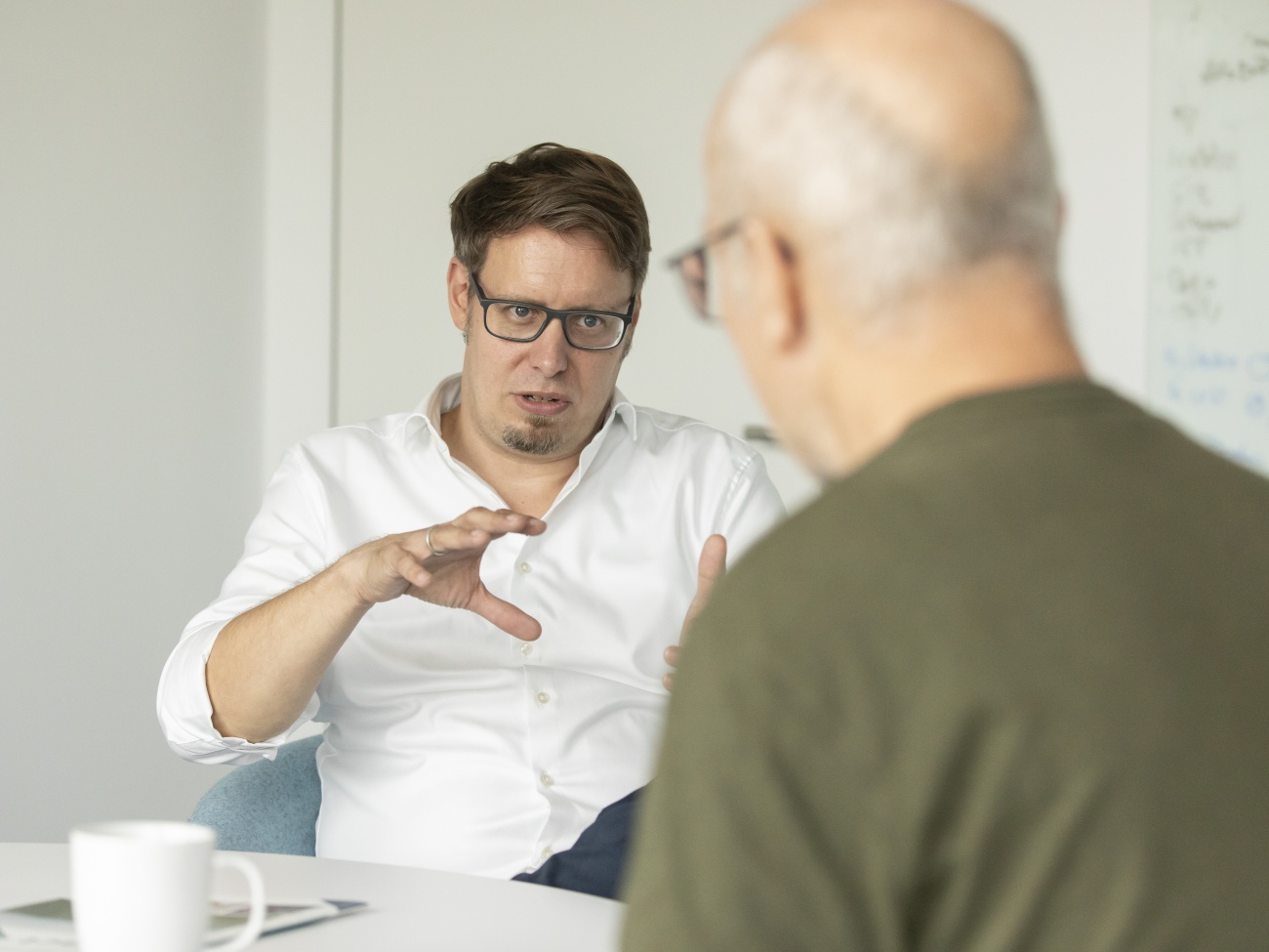 Zwei Männer sitzen an einem Tisch. Der einer ist von vorne zu sehen udn gestikuliert mit den Händen.