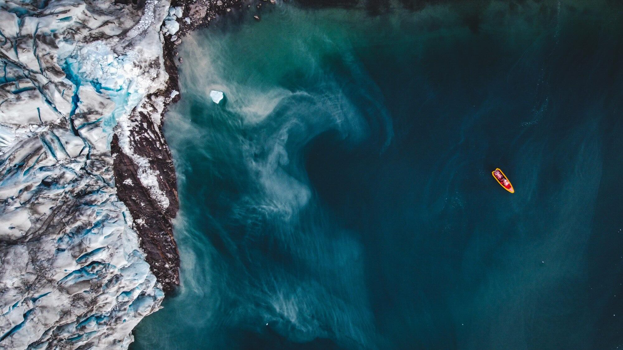 A view from above of the point where a glacier meets the sea, with an orange boat visible in the water. 