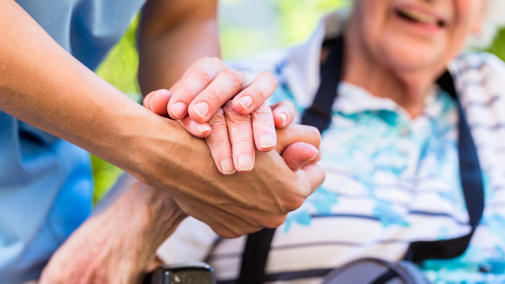 Im Vordergrund hält eine Hand eine ältere Hand. Der Hintergrund ist verschwommen und zeigt Teile der entsprechenden Oberkörper der entsprechenden Personen.