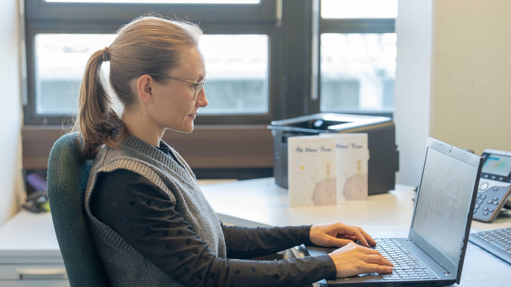 Ms. Hober is typing on a laptop keyboard in an office. In the background, two copies of The Little Prince are leaning against a printer.