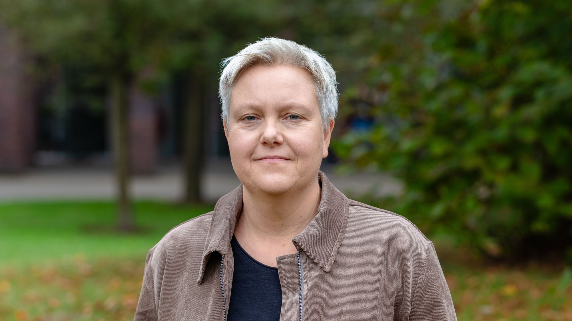 Eine Frau blickt in die Kamera. Im Hintergrund sind verschwommen grüne Bäume zu sehen.