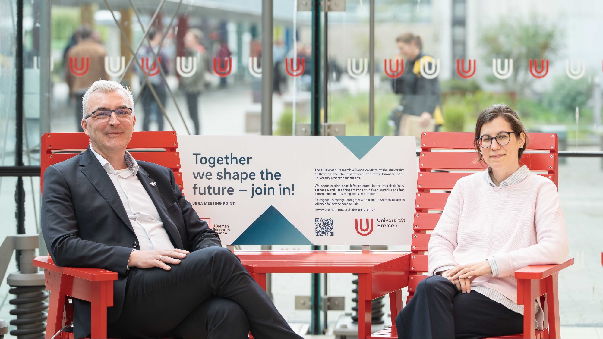 Derk Schönfeld and Jacqueline Schäfer are sitting on a red bench, known as the UBRA Meeting Point, in the Glass Hall on the university campus.