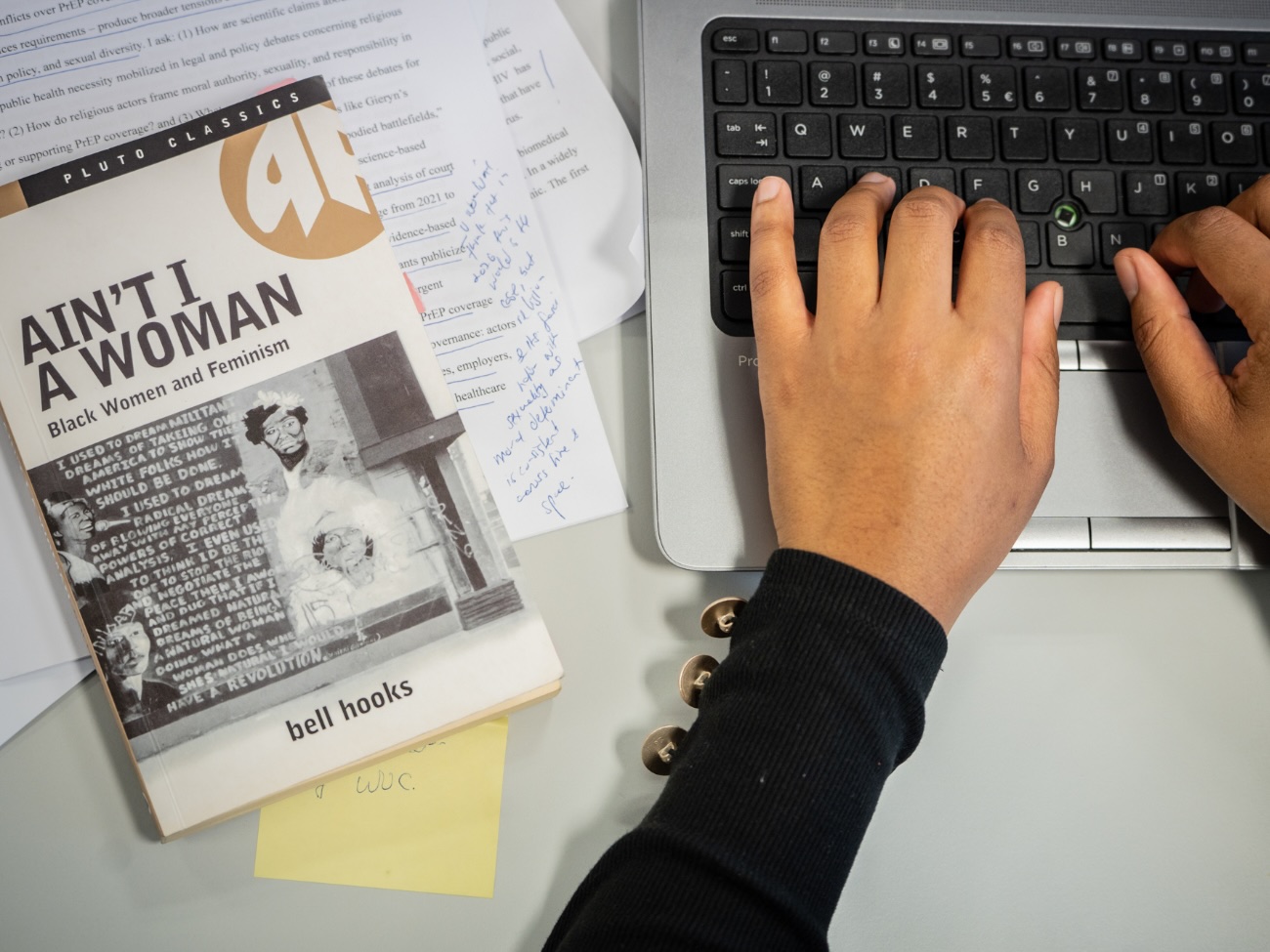Das Bild zeigt einen Blick von oben auf den Ausschnitt eines Schreibtisches. Man sieht darin zwei Hände, die auf eine Laptop-Tastatur tippen. Neben der Tastatur liegt das Buch „Ain’t I a Woman“ von Bell Hooks auf einem kleinen Haufen von annotierten Texte