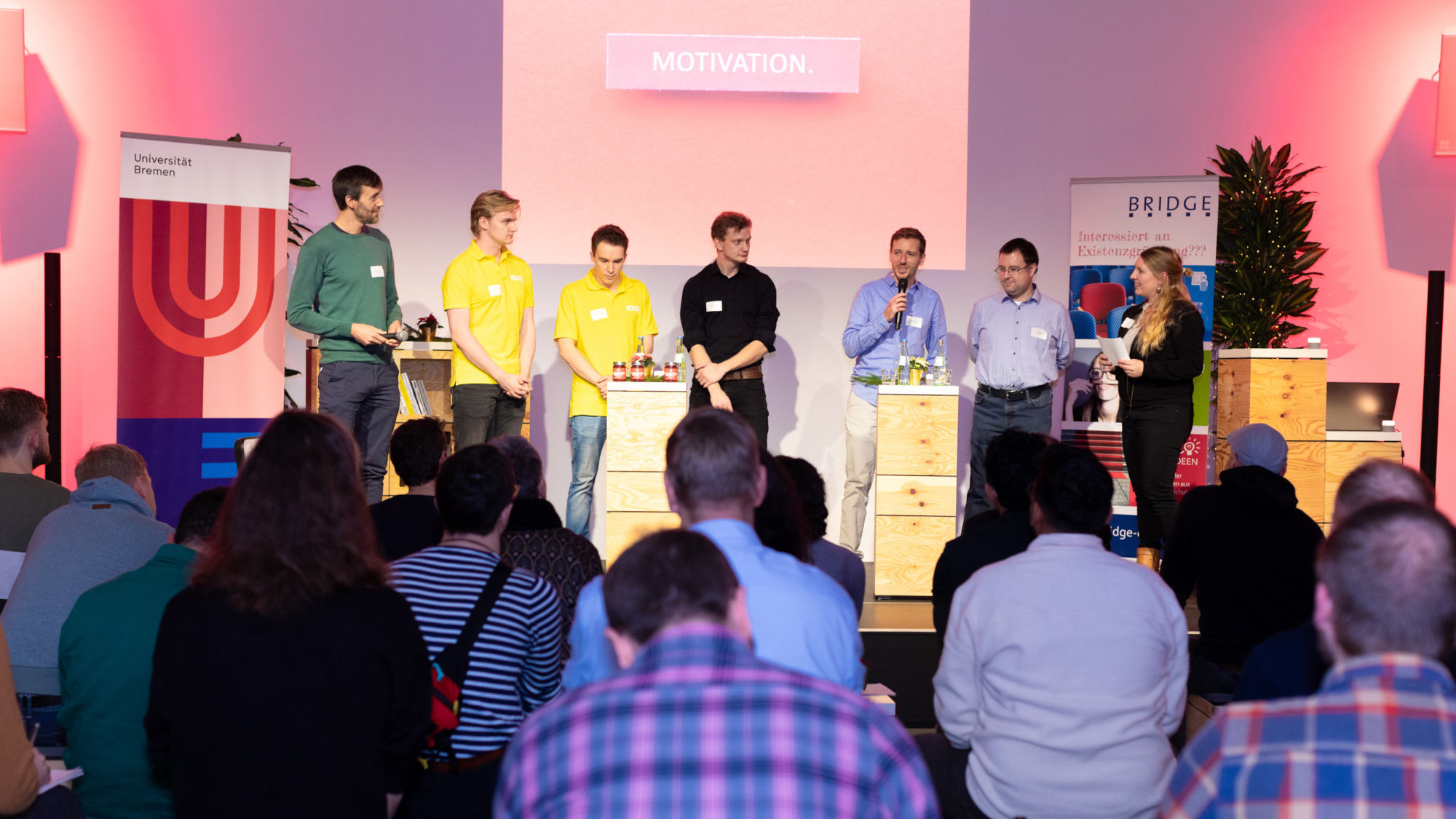 Six men and a woman stand in front of an audience on a stage. A person speaks into a microphone.