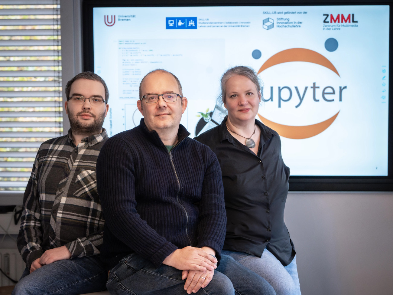 Isabell Schaffer, Marcel Meissner und Heiko Pälike sitzen auf einem Tisch, im Hintergrund ist eine JupyterHub Präsentation zu sehen.