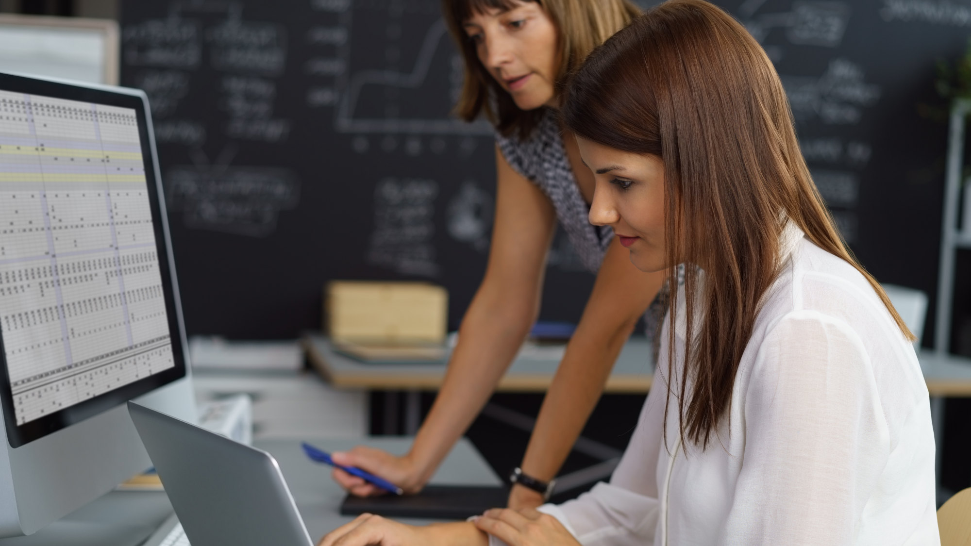 Eine Frau sitzt an einem Schreibtisch vor dem Laptop, eine andere Frau schaut ihr über die Schulter