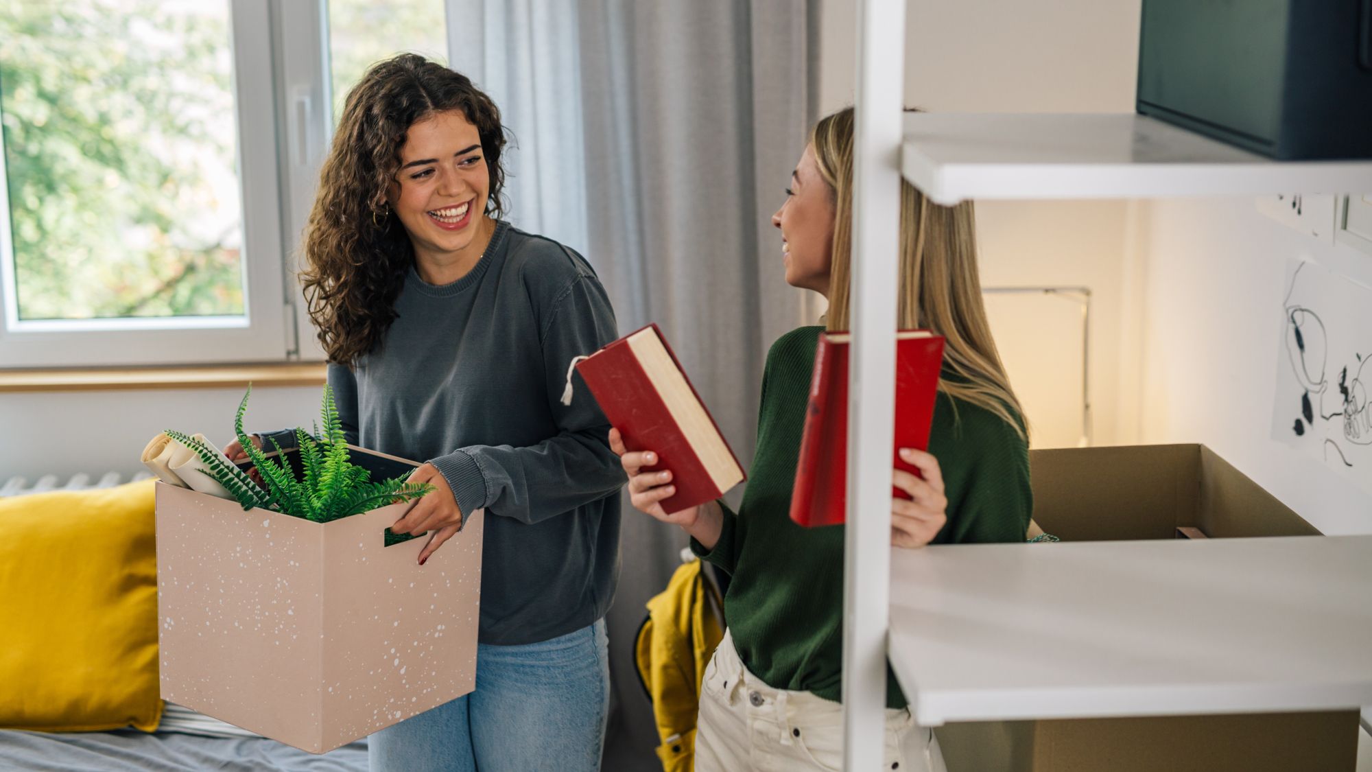 Eine Frau steht mit einem Umzugskarton in einem Raum und lächelt eine weitere Frau mit zwei Büchern in den Händen an.