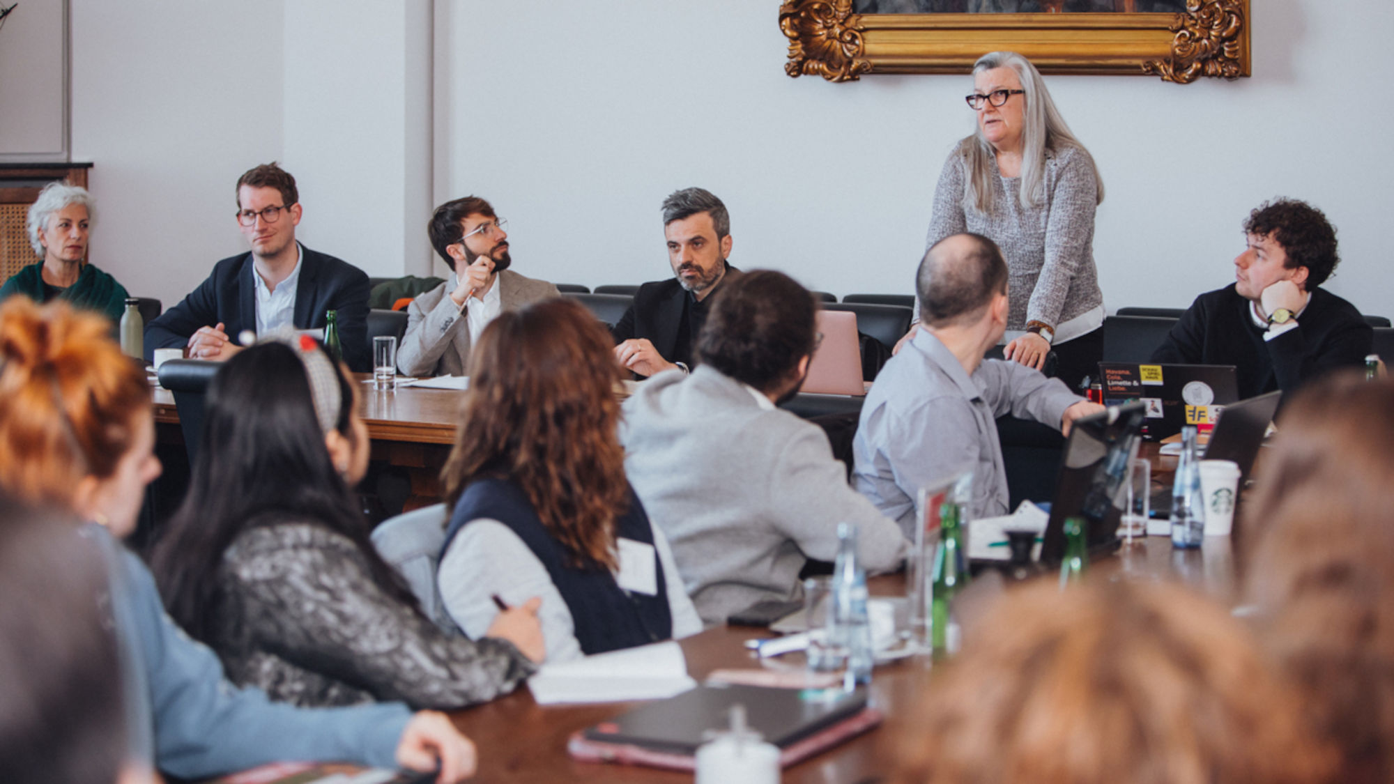 Studierende sitzen an langen Konferenztischen und hören dem Vortrag einer Frau zu.