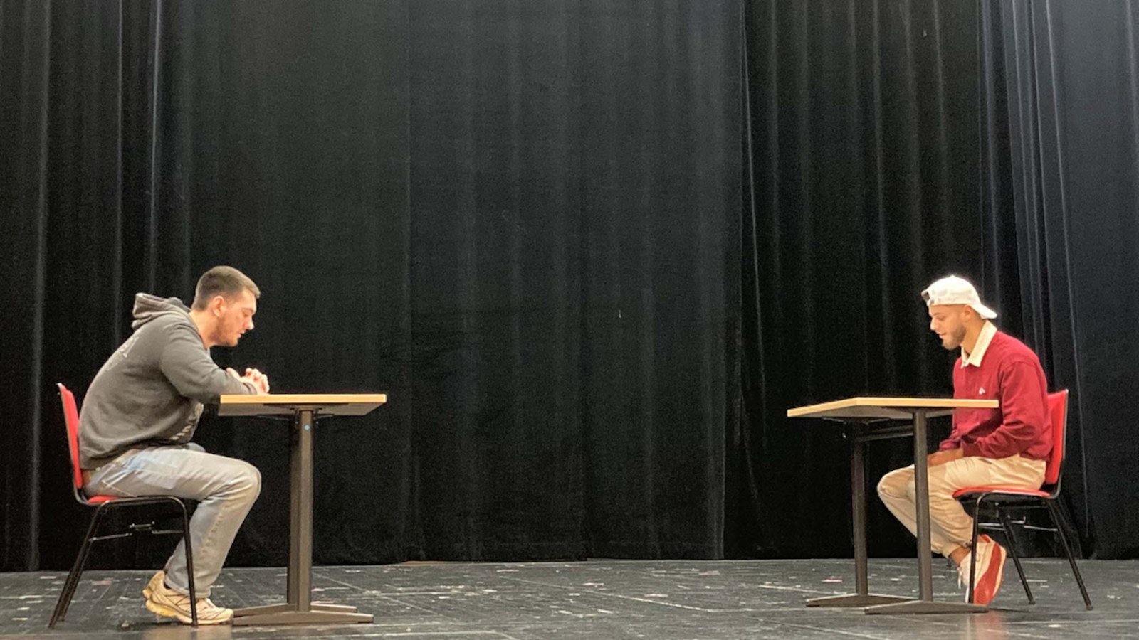 Two young people sit opposite each other on chairs on a stage. Their gaze is fixed on something lying on tables in front of them. 