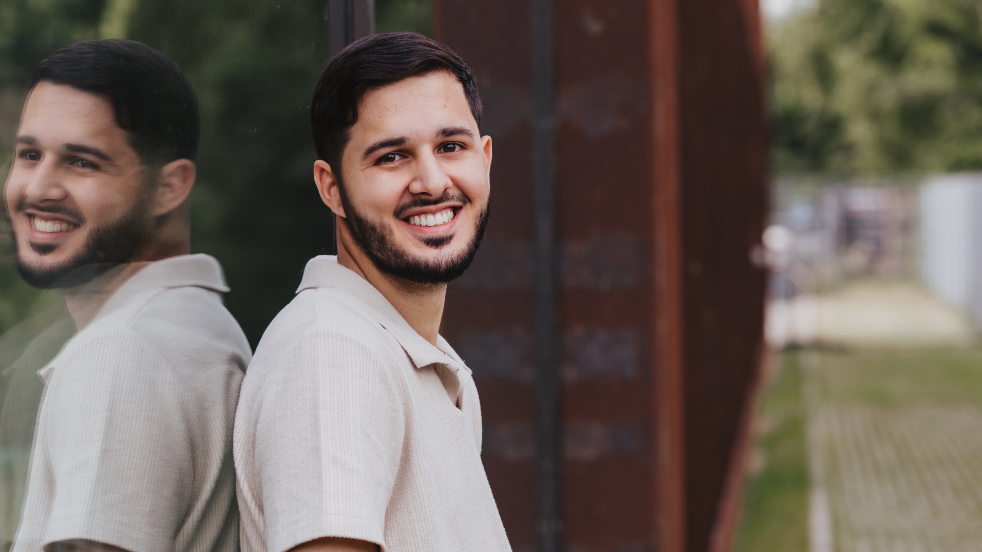 Azer leans against a glass building and smiles at the camera. 