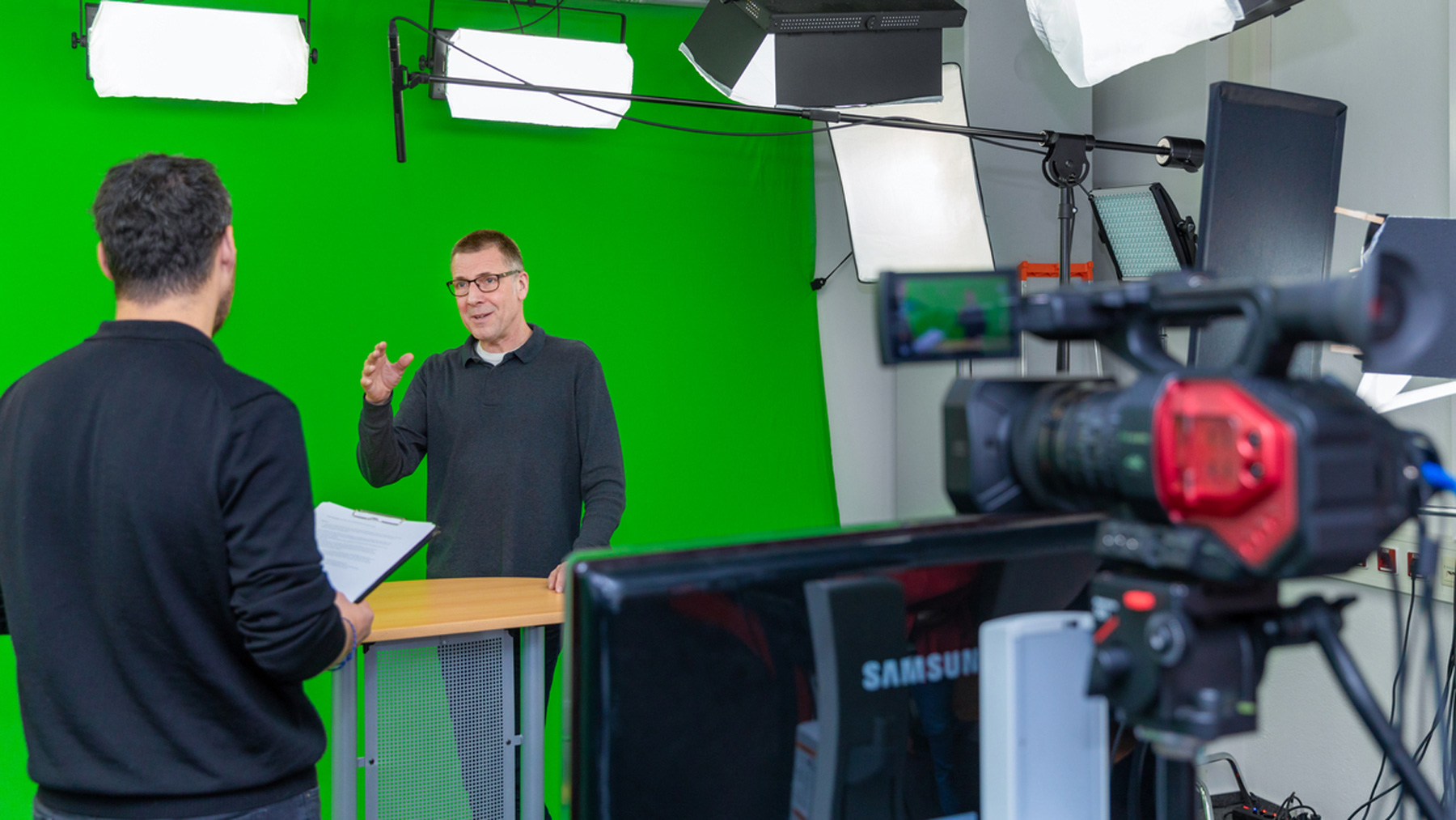 Denis Pijetlovic is standing in front of a greenscreen shooting new learning videos, which can be seen by a camera set up in front of him.