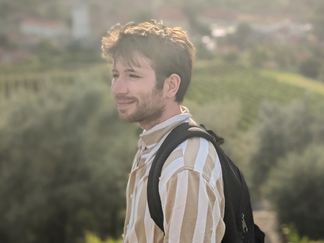 Ein junger Student mit Rucksack steht seitlich vor einem grünen Hügel.