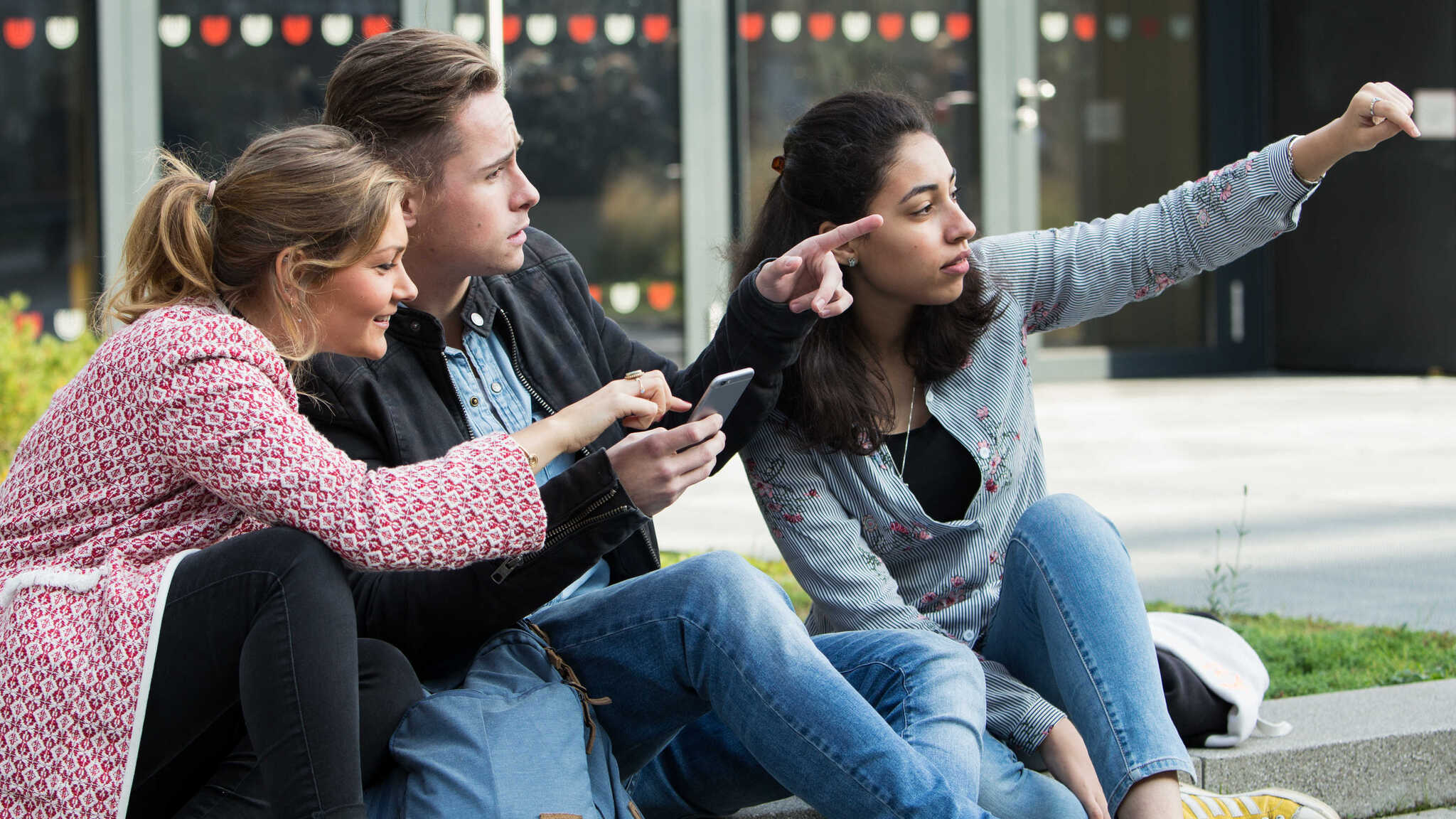 Three students are sitting outside on campus. They are looking in the direction that two of them are pointing.