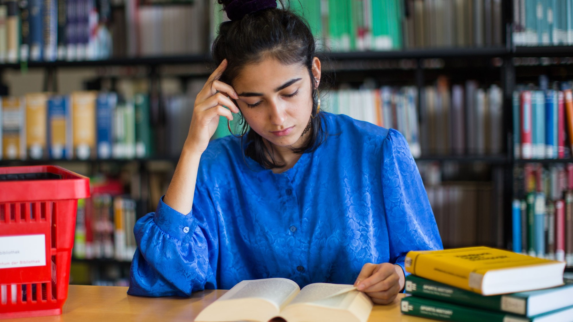 Eine Frau sitzt an einem Tisch und blickt in ein Buch. Auf dem Tisch liegen weitere Bücher und im Hintergrund stehen Bücherregale.