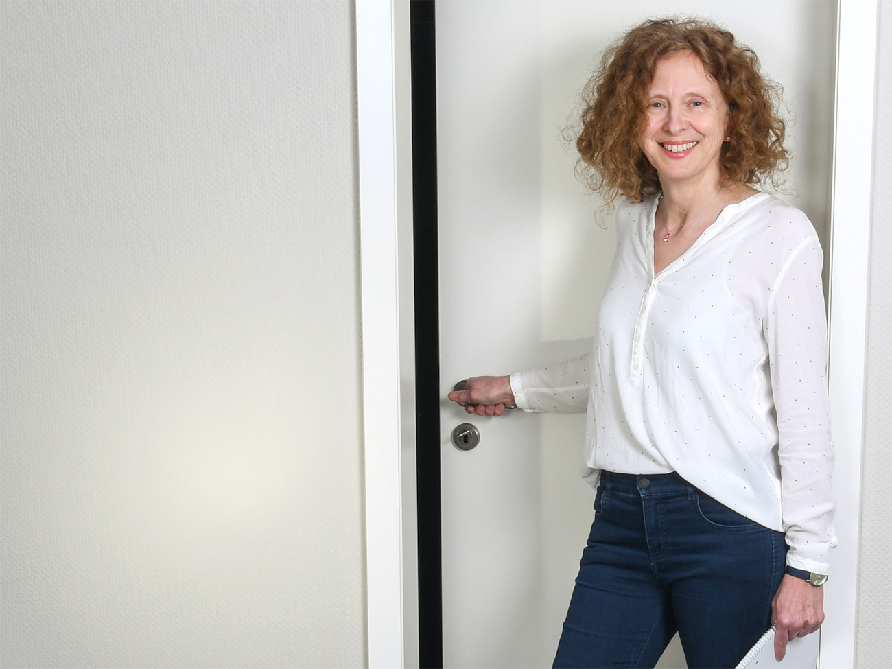 Habilitated university lecturer Iris Stahlke stands in front of a white door. She holds the door handle in her hand and smiles at the camera. 