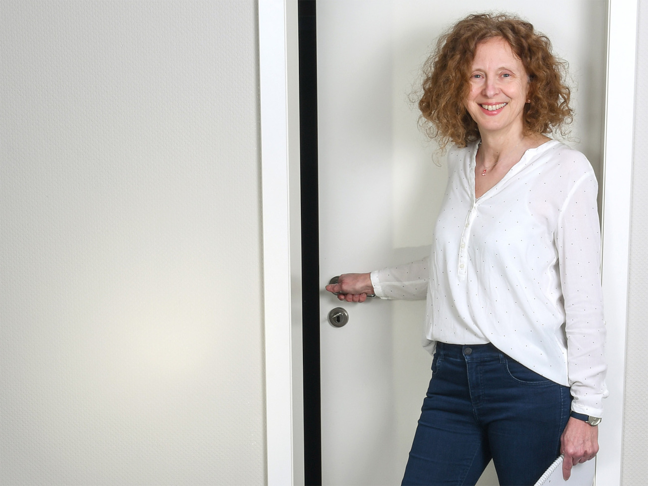 Die habilitierte Universitätslektorin Iris Stahlke steht vor einer weißen Tür. Sie hält die Türklinke in der Hand und schaut lächelnd in die Kamera.