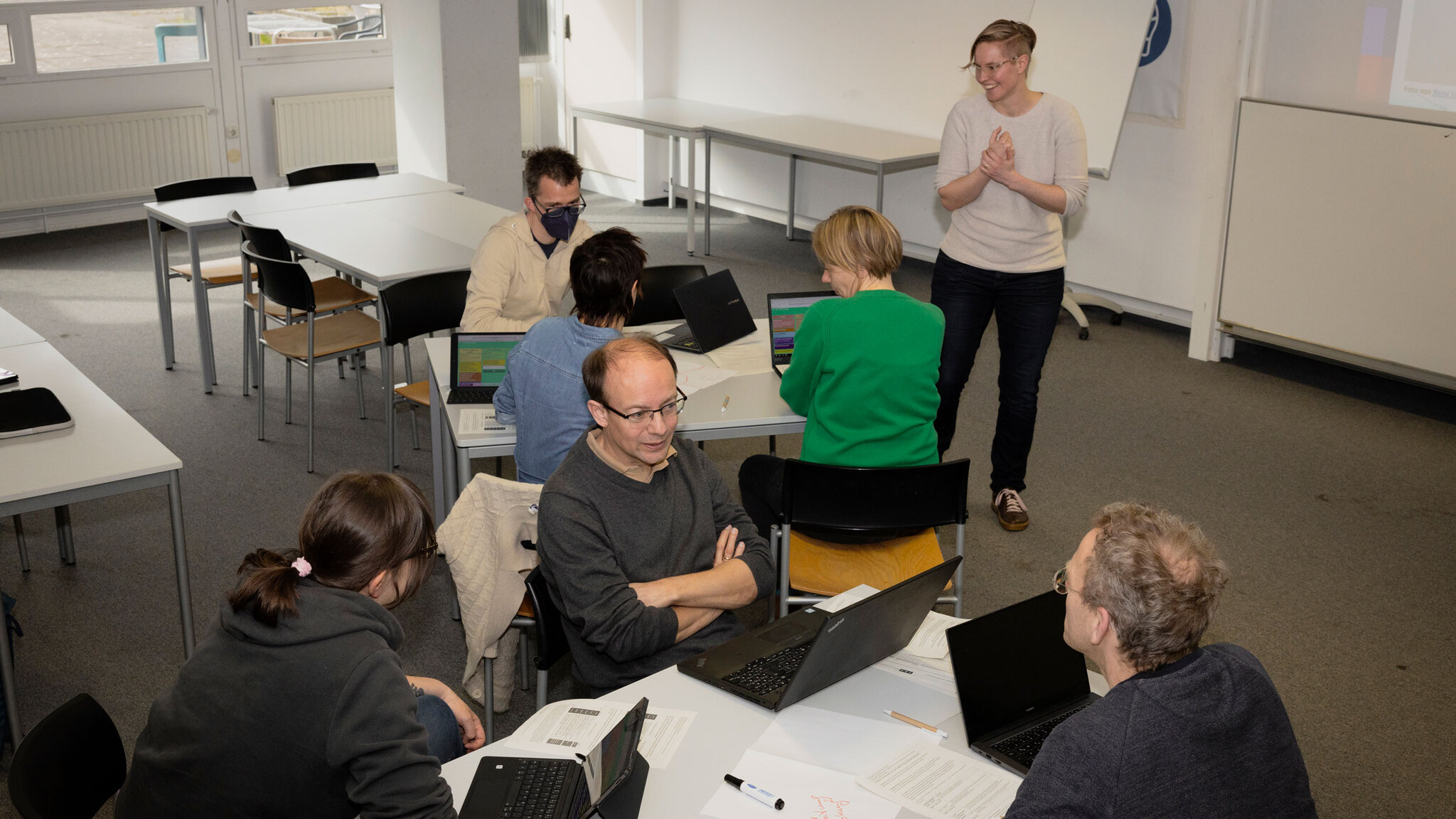 Ein Gruppenarbeitsraum mit mehreren Personen, die an Laptops arbeiten und diskutieren. Eine Person steht vorne, lächelt und scheint eine Präsentation oder Moderation zu leiten. Die Atmosphäre wirkt kooperativ und engagiert. Der Raum ist mit Tischen, Stühl