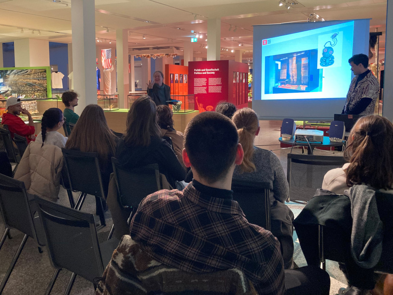 View of the Übersee-Museum in Bremen. Exhibits can be seen in the background. In the foreground, about 20 people sit on chairs listening to a person standing in front of a screen and speaking.