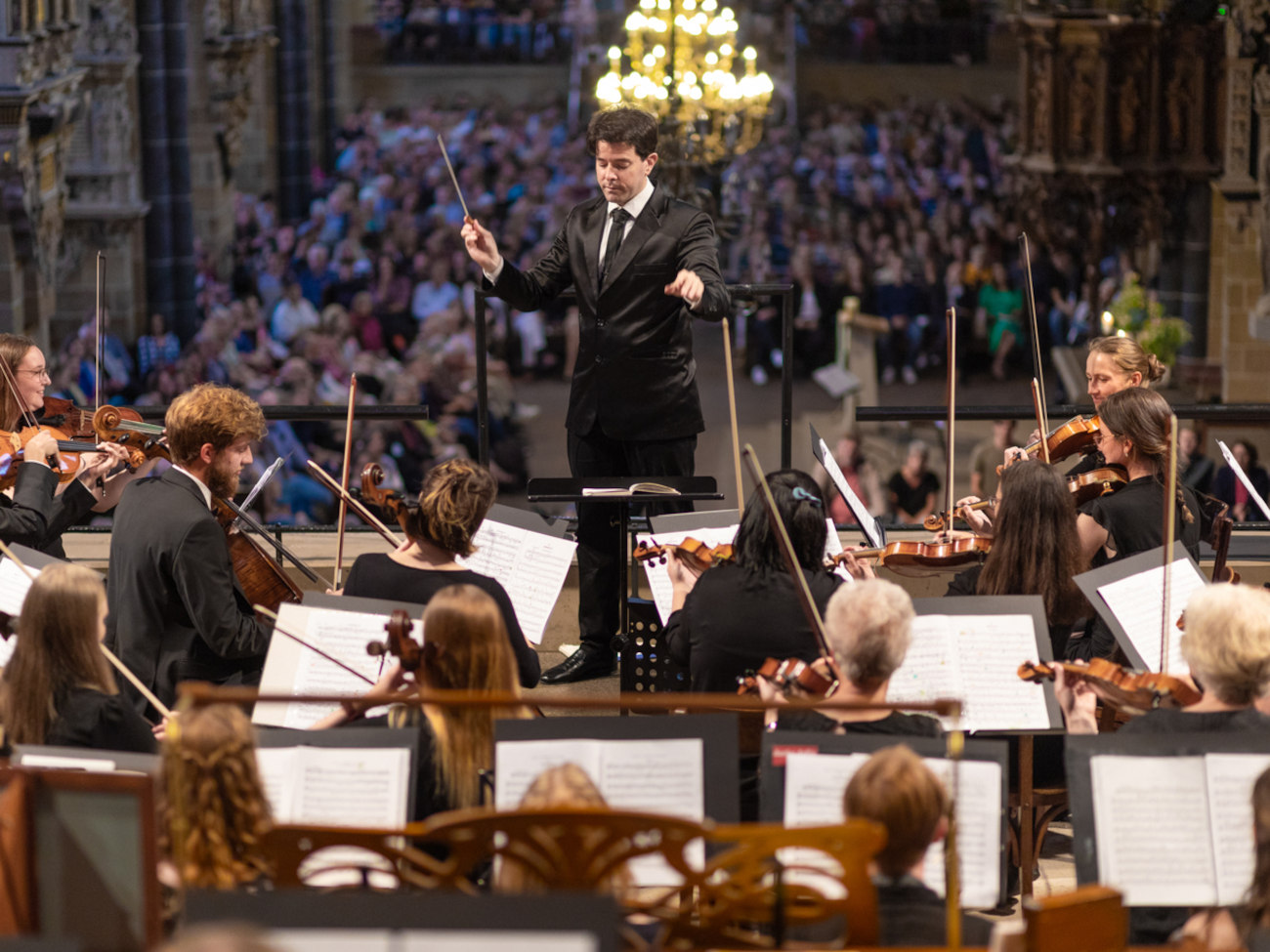 Ein männlicher Dirigent steht vor einem Orchester.