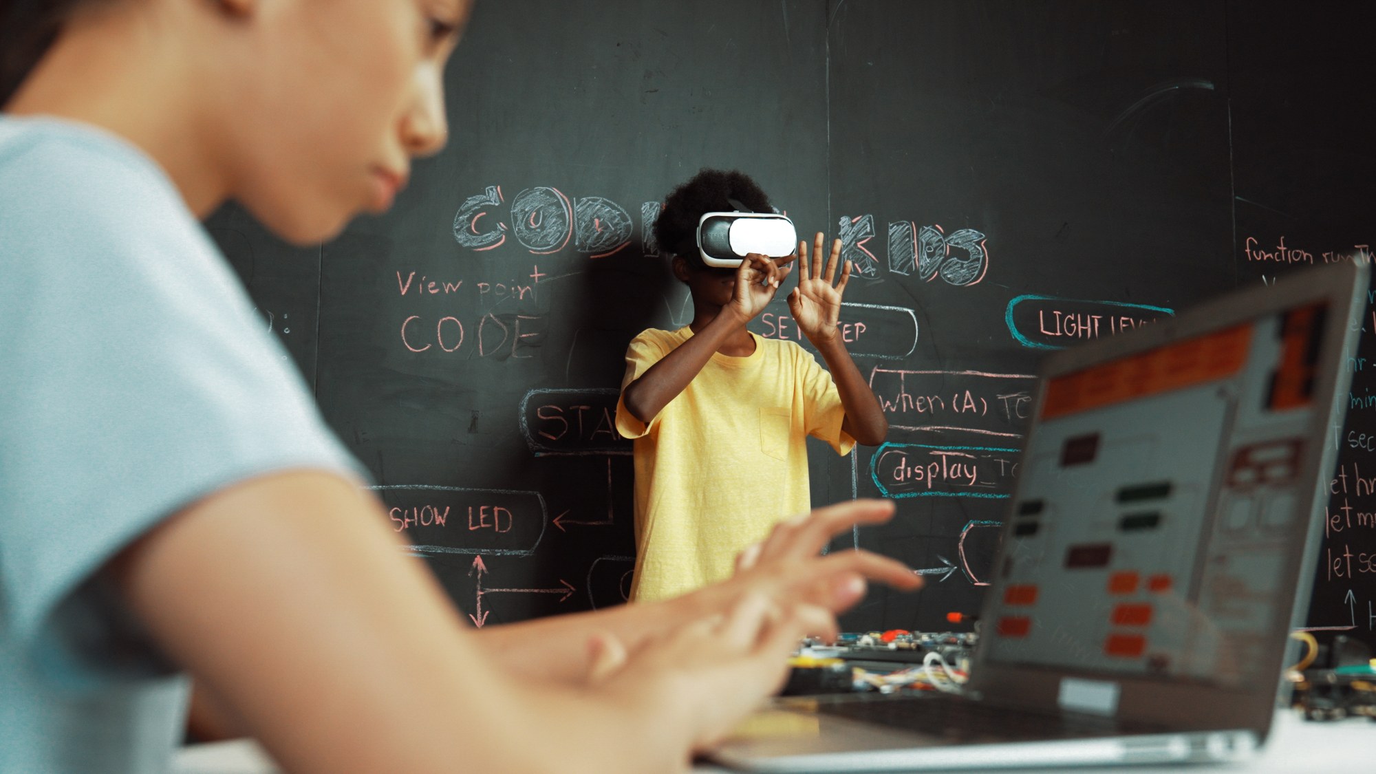 Im Vordergrund sitzt ein Kind an einem Computer. Im Hintergrund steht ein weiteres Kind mit einer VR-Brille.