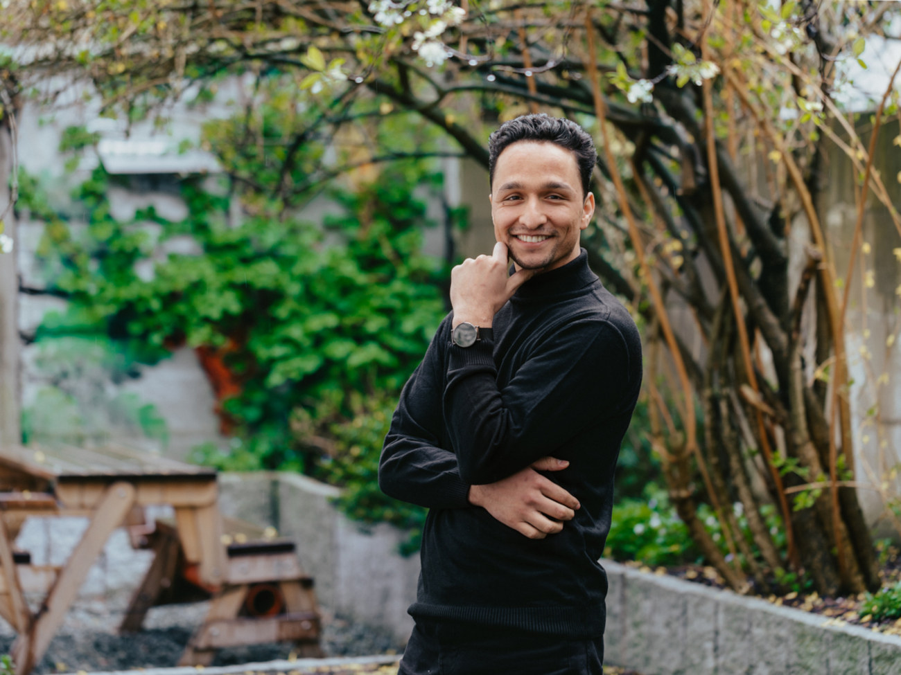 Sohrab Zarbafiyan stands outside and smiles at the camera. A wooden seat can be seen in the background, as well as a small stone wall and a few plants.