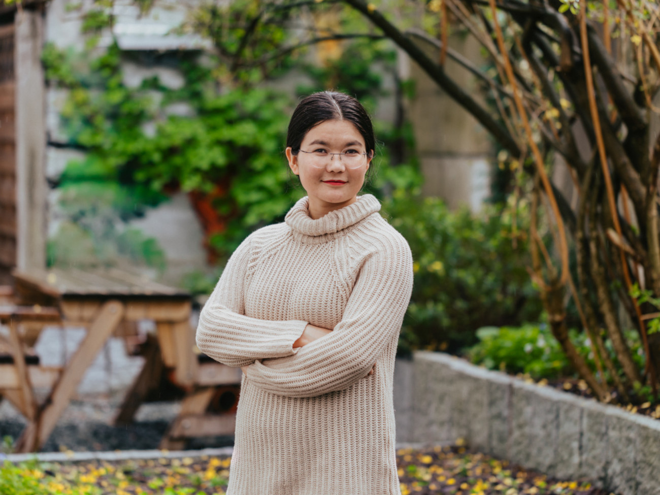 Nazdana Sultanfar stands outside and smiles at the camera. A wooden seat can be seen in the background, as well as a small stone wall and a few plants.