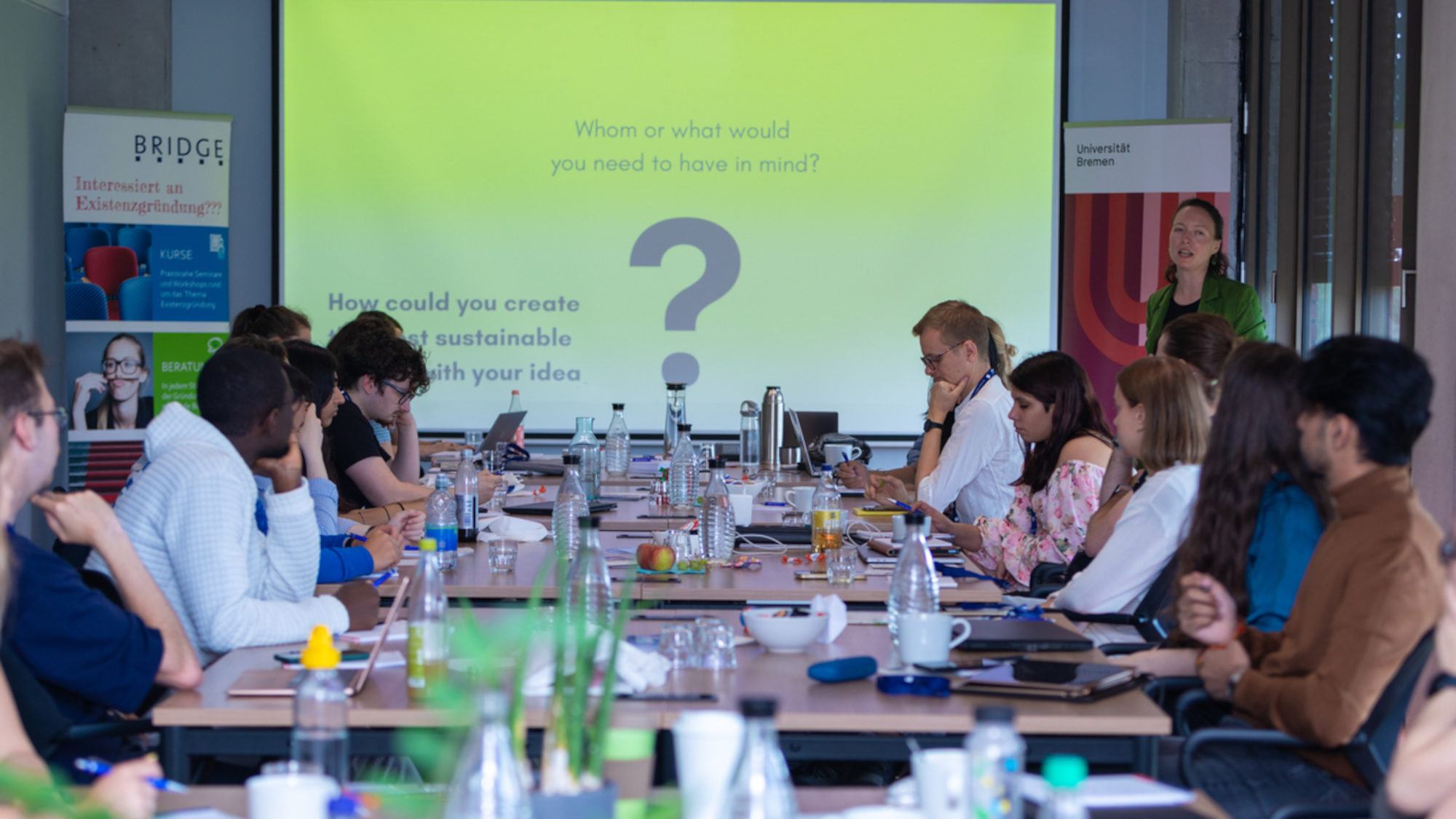 Participants in the Bremen-Cardiff Initiative on Sustainability and Entrepreneurship sit at tables filled with drinks and snacks. A screen can be seen in front, with a person standing next to it.