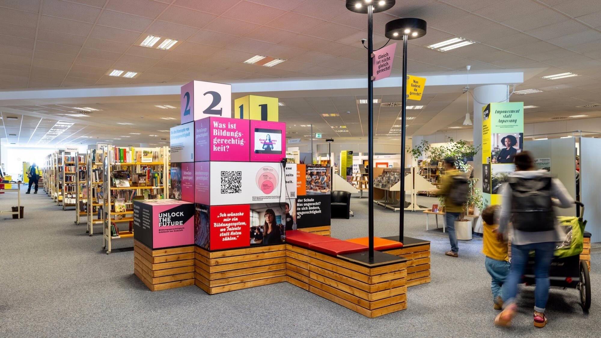 Bibliothek mit Bücherregalen und einem bunten Infostand in der Mitte zum Thema Bildungsgerechtigkeit.
